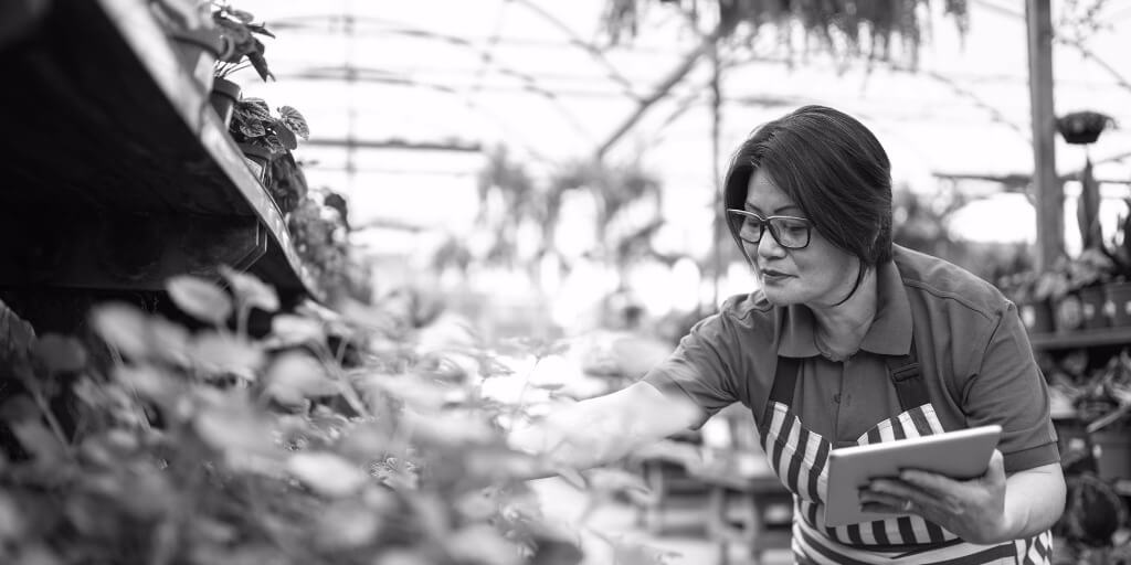 A dark-haired older woman wearing a blue work shirt and a black-and-white striped apron holds a tablet and examines flowers in an outdoor garden center while smiling.