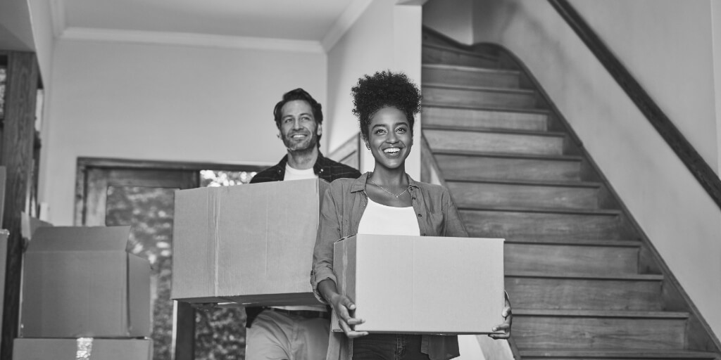 Two people carrying cardboard boxes in a house with a wooden staircase.