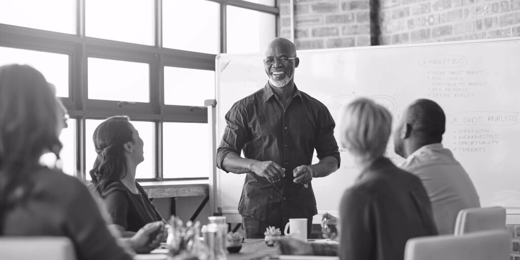 Man leading a team meeting in a modern office, standing and smiling in front of a whiteboard while colleagues sit around a table listening.