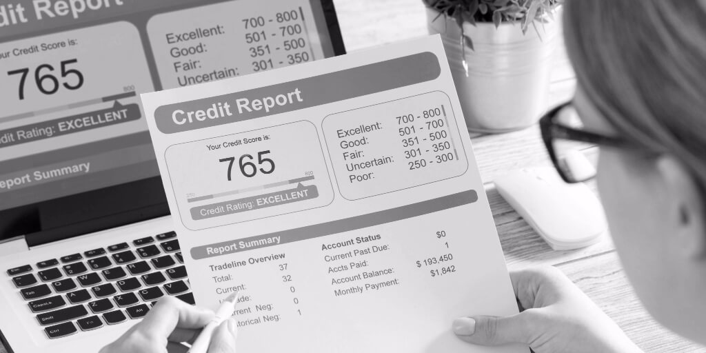 A woman wearing black glasses holds a printed copy of her credit report, which shows that her credit score of 765 is excellent, she has no accounts past due, her current account balance is $193,450, and her monthly payment is $1,842. The same information is shown on the laptop resting on the table in front of her. 