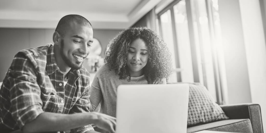 A smiling couple works together on a laptop in a bright, cozy living room.