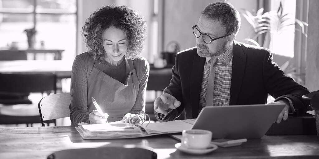 In a sunny dining establishment outfitted with long wooden tables and greenery, a man in a black blazer, blue checkered shirt, silver tie, and black glasses points to a large open binder of paperwork in front of a curly-haired woman wearing an olive green sweater and a red apron as she writes in the binder with a pen.
