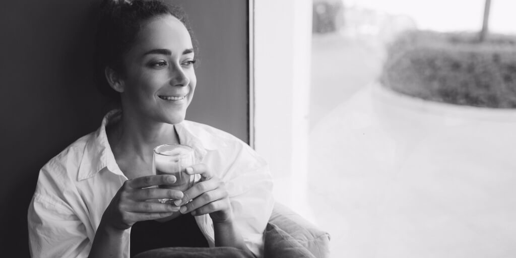 A woman sitting near a window in natural light, coffee in hand, looking thoughtful—not directly at the camera.