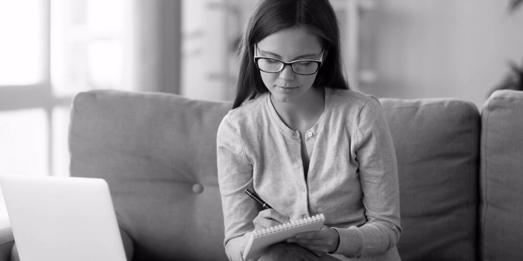 A young woman wearing black and white glasses, a gray long-sleeved shirt, and blue jeans sits on a plush gray couch and writes on a spiral notepad intently, a laptop and two more notebooks resting on the small table in front of her. 