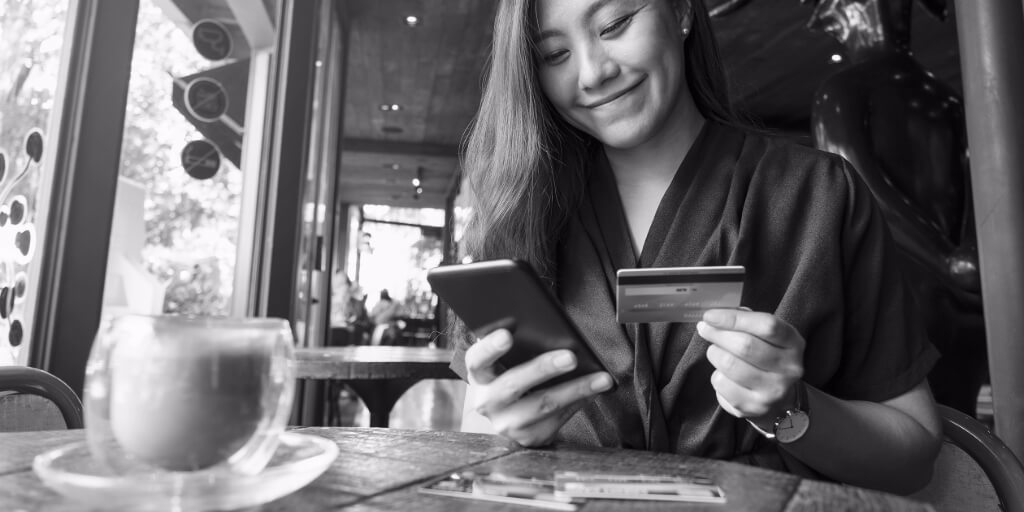 At a rustic wooden round table in a café, a smiling woman in a deep blue blouse holds her cell phone in one hand and a yellow credit or debit card in her other hand.
