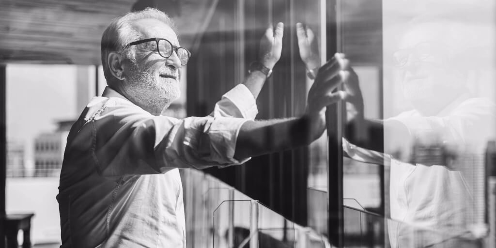 A man with white hair, white facial hair, dark glasses, and a white dress shirt smiles as he looks out over a sunny city through the large glass windows of an office building.