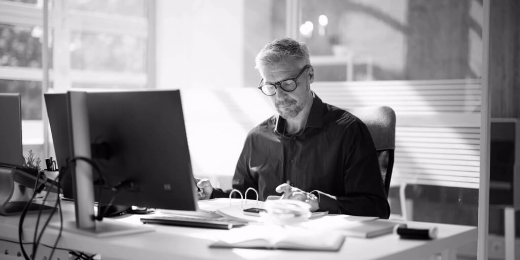 A man with gray hair works at a desk in an office, reviewing documents.  